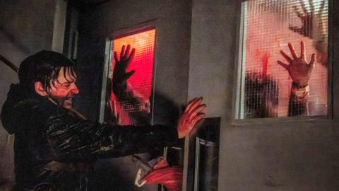 Still from Resident Evil shows Bryan, played by Austin Abrams, desperately pushing against a set of double doors in a hospital hallway. His hands and face are covered in blood, which also matts his hair to his head. A small crowd of zombies can be seen through frosted panes in the doors, their hands pressed against the glass.