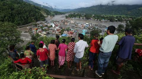 Residents stand on a hillside lookout overlooking a town heavily damaged by flooding. A muddy river cuts through the valley, with debris and sediment covering large parts of the settlement and surrounding riverbanks. Forested mountains rise on both sides under low clouds, while people gather along a railing in the foreground to view the destruction below.