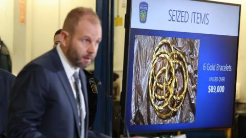 A police officer in a suit stands at a podium and speaks to the media. Next to him is an image on a screen of six gold bracelets with the words "seized items. Valued over $89,000". 