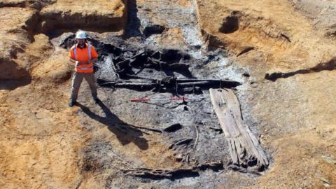 A dug out hole with a big dark patch of soil, where old pieces of wood are protruding in various places, revealing the rough shape of a Middle Bronze Age well. Beside it is a person in a high visibility jacket and wearing a white helmet.