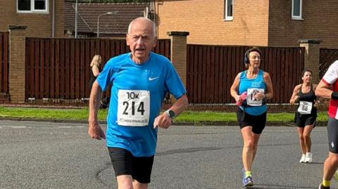 Ian Winship runs in a 10K road race wearing a blue running top and black shorts, with race number 214 pinned to the front, on a residential street in Cumbernauld as other runners follow behind.
