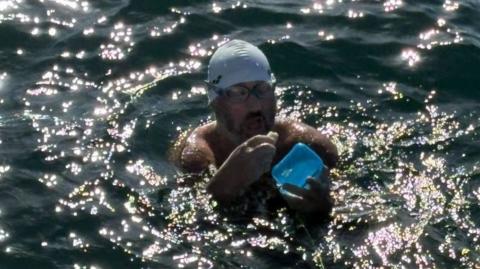 A long-distance swimmer wearing a white swim cap and goggles pauses in open water to eat from a blue container, surrounded by sparkling sunlight reflecting off the sea.