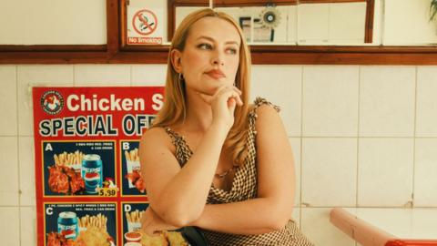 Amelia Dimoldenberg poses in a chicken shop, her chin propped on her right hand and sitting sideways in a traditional booth in front of white tiles and a menu on the wall. She has long blonde hair and blue eyes and wears a strappy checked dress. 