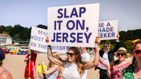 A number of people wearing hats and sunglasses on a beach hold placards reading 'slap it on, Jersey'
