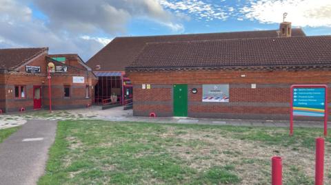 A red brick sports centre building. There is green and brown grass in front of the building and a concrete path.