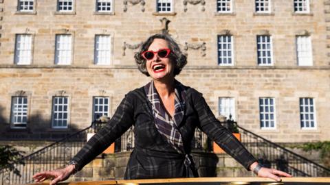 Cathie Boyd stands wearing red cat-eye sunglasses and a dark patterned wrap-style outfit,  behind a circular outdoor installation filled with polished brass bowls. 
She has short curly hair and is leaning forward with both hands resting on the edge of the structure. 
Behind her is Pollok House, with rows of white-framed windows and decorative stonework.