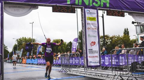 Tim Boswell can be seen at the finishing line of the Worcester half marathon. He is wearing a purple shirt with his race number on the front, and a backpack with a large flag pole sticking out.
