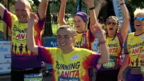 A woman smiling broadly, wearing a yellow t-shirt with red and purple sleeves with the words Running With Anita, has her hands aloft, celebrating. Four other people are behind her, also celebrating and wearing the safe t-shirt. They are outside, a metal fence is in the background. 