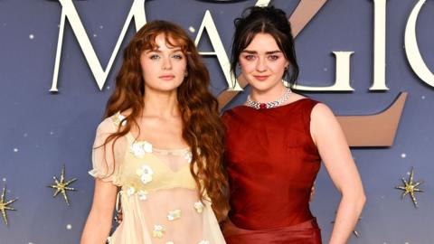 Joey King and Maisie Williams attend the Practical Magic 2 European Premiere in London. Joey wears a cream dress with a lace overlay and flower details and her red hair is worn down in waves. Maisie wears a red dress and her brunette hair in an updo.