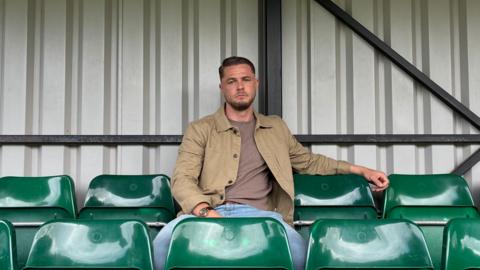 Dan sits on a green chair in a football stand, looking towards the camera. The seats are in a grey metallic stand and Dan wears blue jeans, a maroon top and brown jacket.