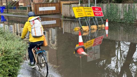 A cyclist splashes through a flooded road ignoring road closure signs and traffic cones