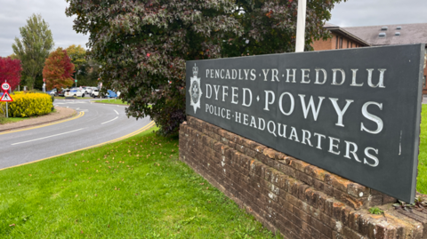 Headquarters of Dyfed-Powys Police - there is a sign that says this on a wall. And a road to the left of the image, and there is a carpark seen towards the back of the image.