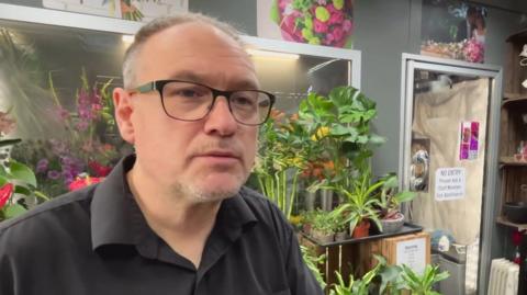 The image shows a person wearing glasses and a dark shirt standing inside what appears to be a florist or plant shop, surrounded by a large number of indoor plants and flowers. Behind the person are shelves displaying pots, bowls and glass vases.