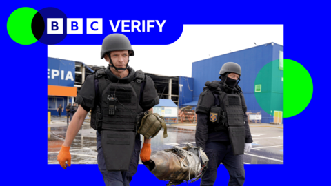 Two Ukrainian first responders carry the remains of a Russian jet-powered drone engine after an attack. They are wearing body armour and helmets and are holding opposite ends of the engine. 