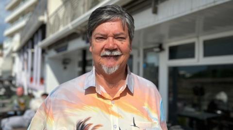 Geoff Mayger is wearing a shirt with palm trees and flamingos on it, and is standing infront of his hotel. He has shirt dark hair and a grey goattee.