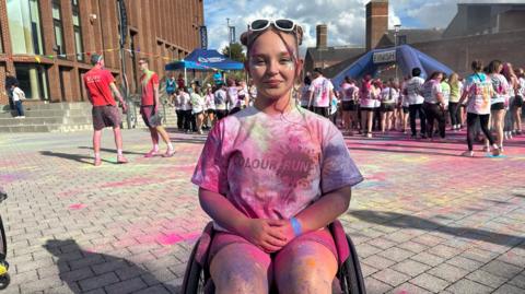 A female university student covered in paint, sat in her wheelchair. She is participating in a fun run event. 