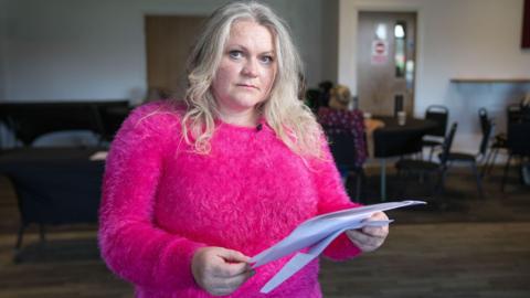 A woman wearing a bright pink jumper holds blood test results in her hand