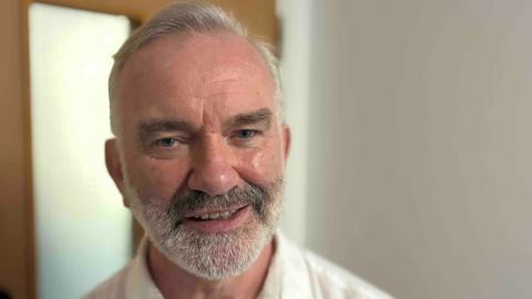 A close-up of the head and shoulders of a man in a white shirt with a white and grey beard and hair. He is smiling at the camera in a white corridor with a door behind him.