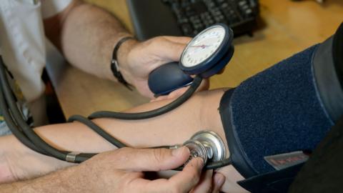 A close up of a doctor holding a stethoscope to a patient's arm. You can only see their ams and not their faces. 
