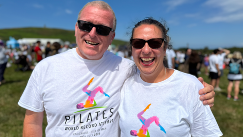 Simon and Karen are standing together in the sunshine smiling wearing white Pilates t-shirts with an illustration of a Pilates move recreating the island's triskelion.