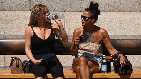 Two women sit on a bench in the sunshine. One woman is taking a sip from a bottle of water, while the other wears sunglasses and holds a fan.
