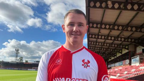 Shaun McEntee wears a red and white Kidderminster Harriers FC jersey. He is stood inside the stadium at the side of he pitch. A stand of red plastic seats are positioned on his left.