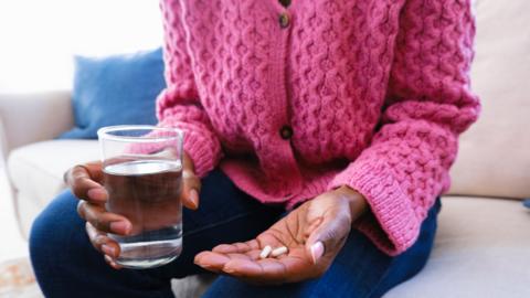 A woman is holding some painkillers in her left hand and glass of water in her right hand. She is wearing a pink cardigan but we cant see her face