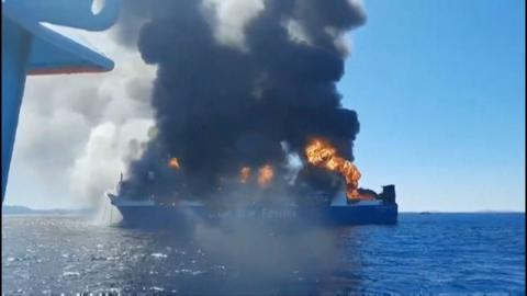 Smoke and flames rise from a cargo ship in the ocean.