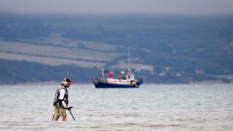A man in the foreground stands in the sea up to his knees with a metal detector and  a fishing boat is seen in the background. The sky is grey and cloudy and the sea looks slightly choppy