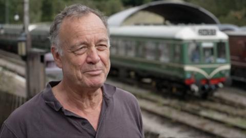 Man in brown top looks past camera while train is seen in the background.