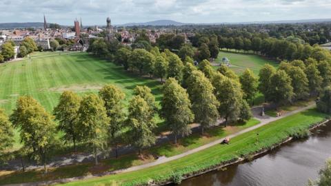 An aerial image of two large green fields spilt up by an avenue of trees. It is bordered by more trees, and in the distance is the town of Shrewsbury. In the foreground there is a river