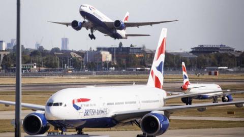 Three British Airways aircraft at Heathrow Airport, one is taking off in the distance with London skyline in the background