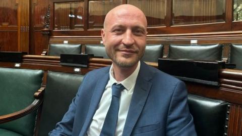 A man sat in a council chamber wearing a blue shirt and tie with a white shirt. The seats are green leather with wooden panels in the background. 