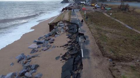 Large chunks of a road have broken after being damaged in a storm. The road is next to a beach.