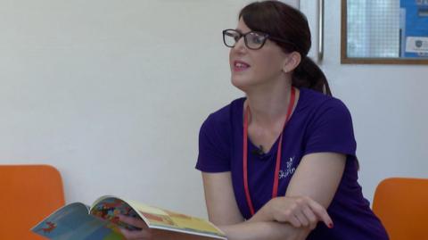 Mariam Badavi, wearing black framed glasses and a purple t-shirt, is sitting on an orange chair in an assembly room with her book in her hand as she looks out to the children who are asking her questions.