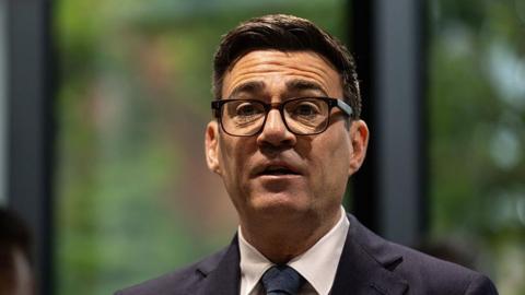 A picture of Andy Burnham from the shoulders up. He is wearing a dark suit with a white shirt and dark tie, has thick-rimmed black glasses on, and has short, dark brown hair. The background behind him is blurred and he is looking forward with his mouth slightly open and his eyebrows raised.