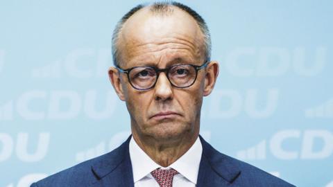 Friedrich Merz stands in front of a party poster giving a broadcast. He wears a blue suit, glasses, red spotted tie, and a sad expression on his face.