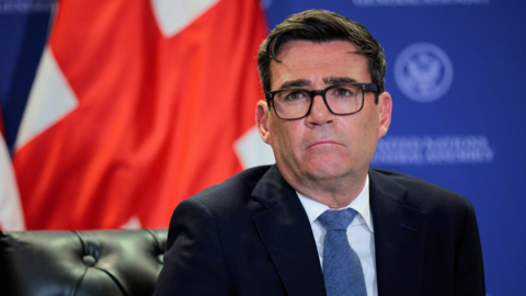 Andy Burnham is wearing glasses and a black suit with a white shirt and a blue time. A blue backdrop is behind him and flags. 