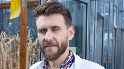 Dr John Ferguson is wearing a white lab coat over a checked shirt. He has brown hair and a beard.