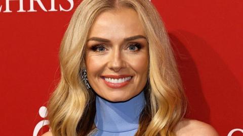 Katherine Jenkins, who has long blonde wavy hair, smiles at the camera. She is wearing a blue halter neck dress and crystal earrings