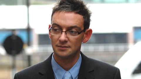 Colin Norris - Portrait photograph of a person wearing glasses, a dark suit jacket and a light blue shirt and tie, standing indoors with a blurred background.