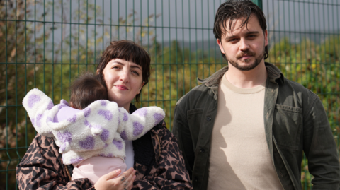 Nel Byers, her fiancé Otis Proctor and their three month old daughter Wren in front of a green fence