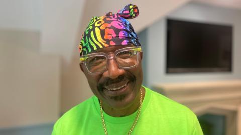 Mr Motivator, who is weaing a bright green t-shirt and a multi-coloured bandana. He also wears a gold chain. He is smiling in the photo.