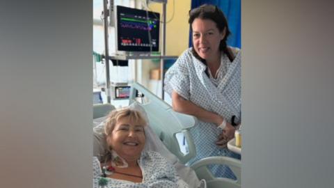 Two smiling women in hospital gowns pose beside a hospital bed and vital signs monitor.