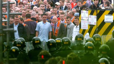 A group of police in riot gear are facing a large crowd of people. Mostly are men.  Many are wearing orange sashes. Signs say decommission the parades commission. 