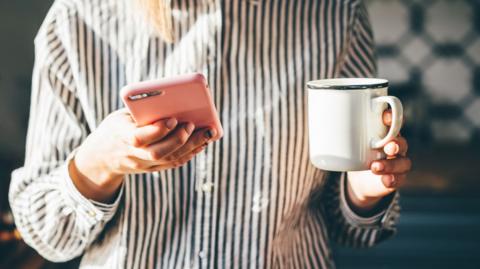 A woman in a striped shirt holds her phone, which has a pink cover. In the other hand she holds a cup of tea. Her face is not visible. 