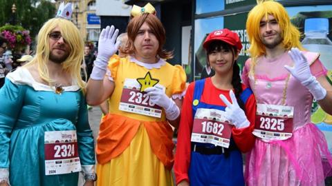 Four people, dressed up in fancy dress, the man on the left has on a blonde wig, and blue dress, the next man is wearing a brown wig and yellow dress, a woman is dressed up as a Super Mario character, and a man, on the right is wearing a blonde wig and pink dress. Three of them are waving.