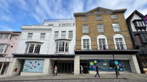 A white building with pillars on its front and large shop windows at the ground floor. Above there are more floors with windows and a balcony.