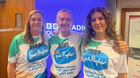 Dave Cant pictured in the middle of his daughter Debbie, who is on the left, and his granddaughter Issy, on the right. They are standing in front of a BBC Radio Gloucester sign. All three are wearing white t-shirts with the Sue Ryder logo on the front.