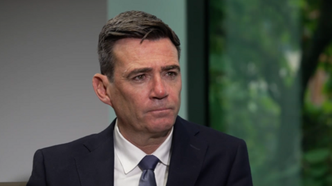 A head and shoulders shot of Prime Minister Andy Burnham being interviewed. He is looking off to the right of the photo. He has black hair and is wearing a dark suit jacket over a white shirt, with a dark blue tie. Behind him is a window with greenery outside. 